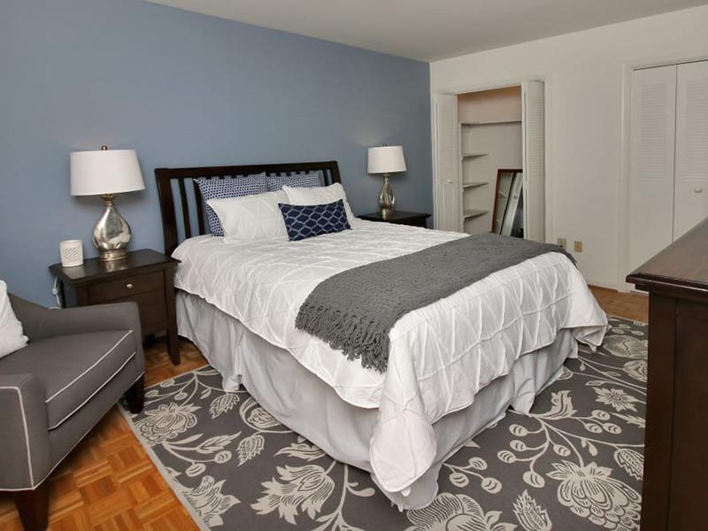 The apartment bedroom is furnished with a large bed, nightstand and upholstered chair with a walk-in closet.